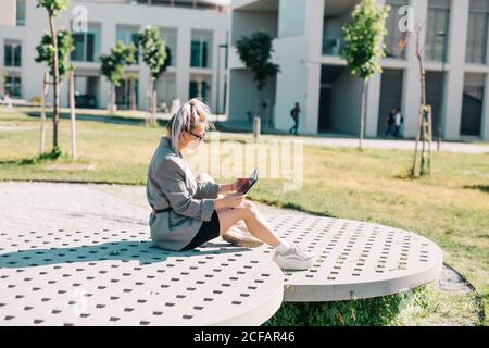 Side view of trendy businessWoman working outside sitting on round decorative panel plates nearby building in bright day Stock Photo