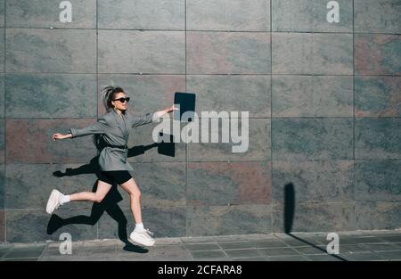 Side view of stylish businessWoman active jumping along marble wall and waving hands with tablet leaving contrast shadow in light day Stock Photo