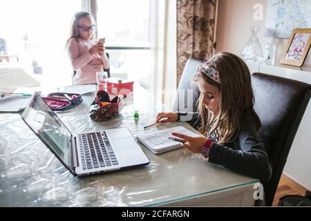 Smart girl taking notes in notebook while sitting at table near sister and doing homework task from laptop at home Stock Photo
