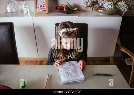 From above focused girl sitting at table and reading notes in notepad while doing homework assignment at home Stock Photo