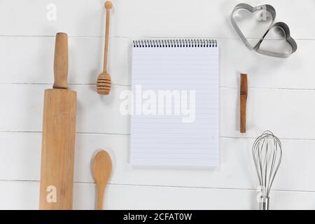 View of a composition with a notebook with cookie cutters, whisker and rolling pin on a white wooden Stock Photo