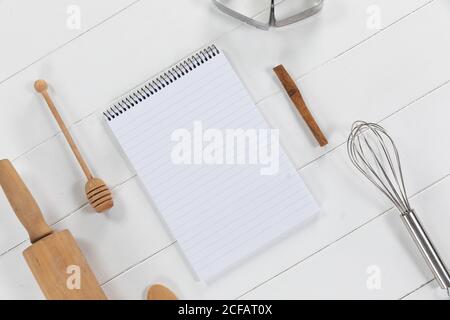 View of a composition with a notebook with cookie cutters, whisker and rolling pin on a white wooden Stock Photo