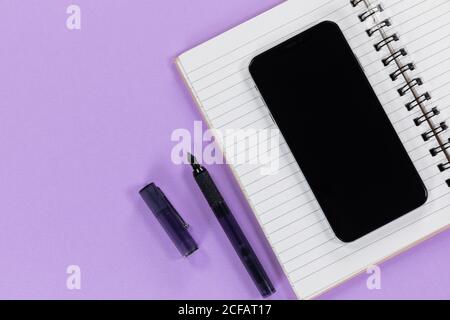 View of a black smartphone, a notebook and a black pen on plain purple background Stock Photo