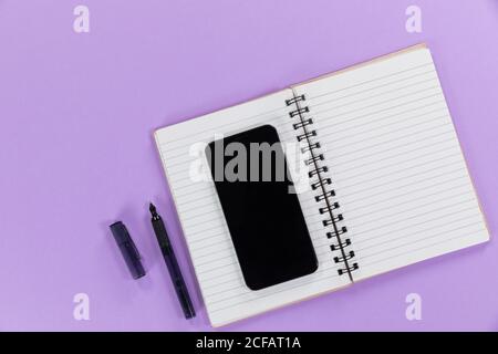 View of a black smartphone, a notebook and a black pen on plain purple background Stock Photo