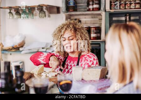 Happy adult Woman with curly hair talking with customer while selling cheese in local food delicatessen store Stock Photo