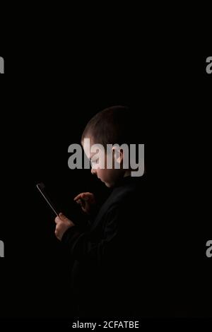 Adorable serious child in casual wear holding tablet in hands with determined look isolated on black background Stock Photo