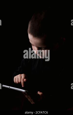 Adorable serious child in casual wear holding tablet in hands with determined look isolated on black background Stock Photo