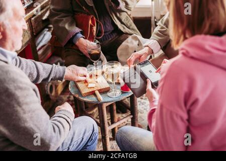 From above crop Woman using smartphone while sharing company with elderly men tasting cheese and wine in cozy local food shop Stock Photo