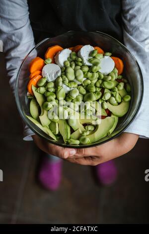 Hands of anonymous little girl holding bowl of healthy vegetable salad while standing in kitchen at home Stock Photo