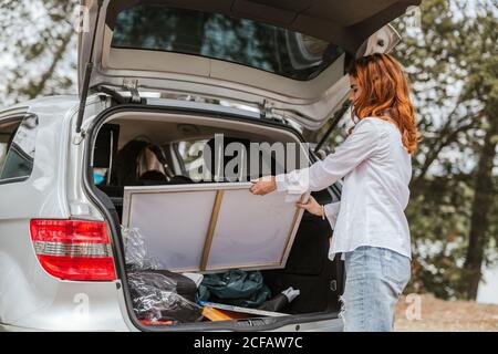 Side view of young Woman in casual outfit taking canvas from open trunk of modern car in countryside Stock Photo