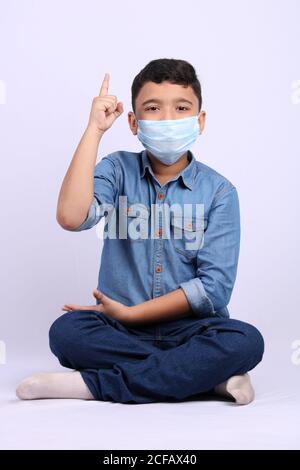 indian cute young boy wearing surgical mask pointing finger towards top while seating on floor. Stock Photo