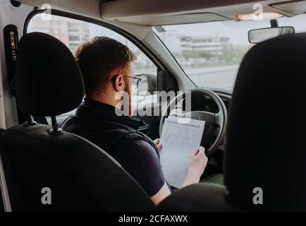 Back view of attentive thoughtful adult man focusing and checking documents while sitting behind steering wheel in car cabin during daytime on blurred background Stock Photo