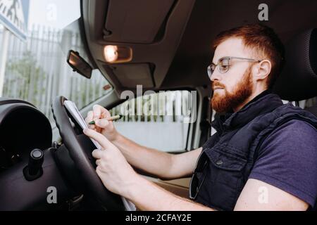 Side view of attentive thoughtful adult man focusing and checking documents while sitting behind steering wheel in car cabin during daytime on blurred background Stock Photo