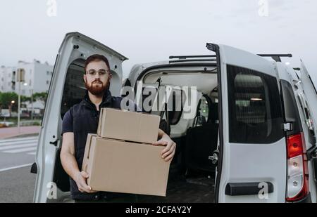 Attentive adult bearded courier in glasses carefully unloading cardboard boxes from car for further delivery to customer on blurred background during daytime Stock Photo