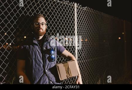 Calm confident adult bearded courier in glasses with boxes looking at camera while standing and leaning on mesh fence on street at night Stock Photo