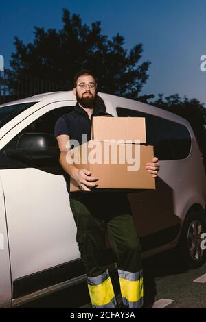 Adult bearded man in glasses thinking and looking at camera while standing near car and holding cardboard boxes in evening Stock Photo