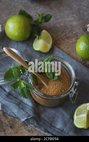 Overhead brown sugar in a jar near fresh limes and peppermint leaves placed on napkin on a wooden table Stock Photo