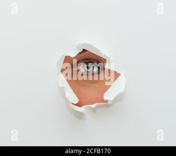 Blue eye of unrecognizable female looking at camera through ripped hole in white paper cardboard background Stock Photo