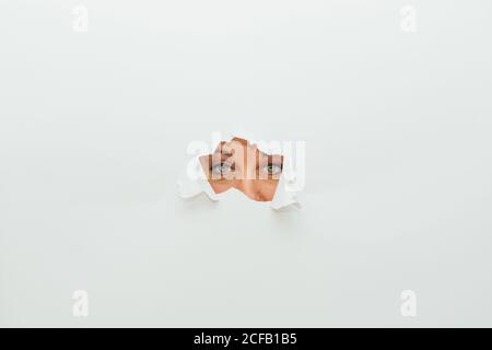 Blue eyes of unrecognizable female looking at camera through ripped hole in white paper cardboard background Stock Photo