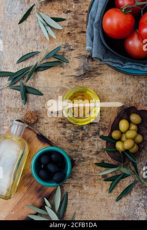 From above of glass jar with organic olive oil with wooden spoon placed on shabby gray table with fresh tomatoes green and black olives near tree branches Stock Photo