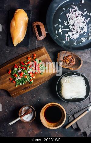 Top view of uncooked rice noodles in bowl placed among ingredients for recipe including chicken fillet and chopped vegetables with soy sauce and spices Stock Photo