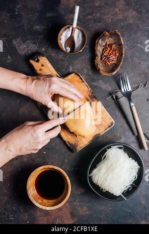 From above of crop anonymous Woman cutting raw chicken fillet on wooden cutting board while preparing noodles for lunch Stock Photo