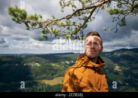 Portrait of cheerful traveler. Young man under branch of tree looking at view. Stock Photo