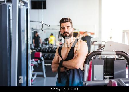 Muscular bearded man in sportswear looking at camera while standing with arms crossed and leaning on sports equipment in light contemporary gym Stock Photo