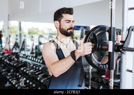 Muscular bearded man in sportswear standing near squat rack and putting weight disk on barbell while preparing for weightlifting training in modern gym Stock Photo