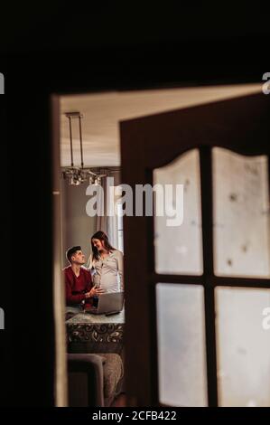 Young man in red jumper sitting on couch and kissing holding wife standing nearby while spending free time at home and enjoying time together Stock Photo