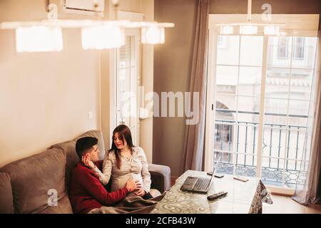 Young man in red jumper sitting with pregnant wife in beige blouse on sofa and touching baby bump while spending together on the computer winter day at home Stock Photo