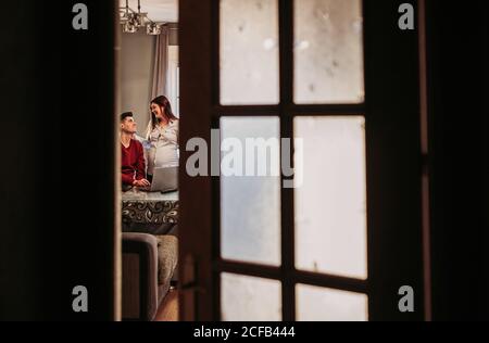 Young man in red jumper sitting on couch and kissing holding wife standing nearby while spending free time at home and enjoying time together Stock Photo