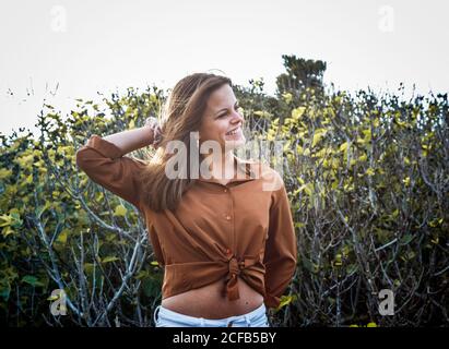 Cheerful beautiful Woman standing in front of bushes touching back of head with hand looking away Stock Photo