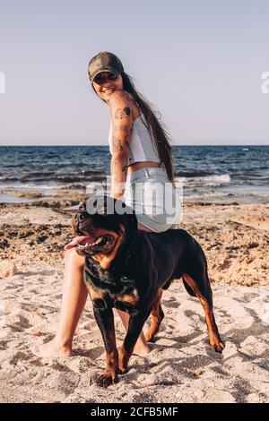 Positive young lady looking at camera in casual clothes and cap standing on sandy beach with Rottweiler dog while spending time on sunny day in summer Stock Photo
