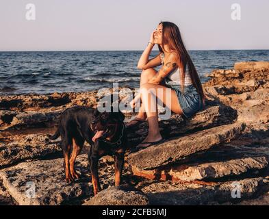 Side view of trendy tattooed Woman looking away while sitting on stone with Rottweiler dog while spending time on rocky seashore Stock Photo