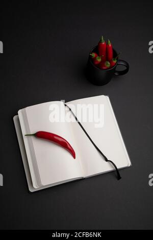Mug of hot red chili peppers placed on black background near open notepad with blank pages Stock Photo