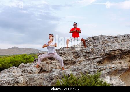 Adult man and Woman with closed eyes and clasped hands standing on rough stone and meditating while practicing Tai Chi against overcast sky Stock Photo