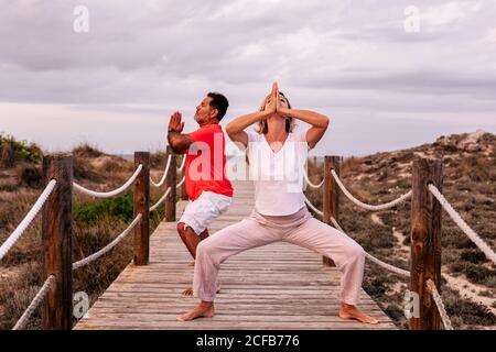 Full body barefoot man and Woman clasping hands and meditating while standing on lumber path against overcast sky in nature Stock Photo