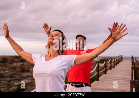 Adult man and Woman stretching out arms and breathing while standing on wooden path against overcast sky Stock Photo