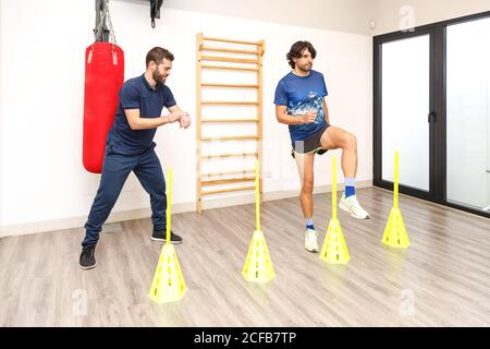 Young man in active wear standing on one leg while doing exercise with yellow cones on speed with trainer smiling and checking time on wristwatch in gym Stock Photo