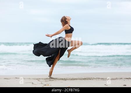Side view of beautiful attractive female in black outfit dancing on sand near waving sea Stock Photo