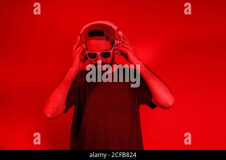 Stylish bearded guy in hat and sunglasses holding headphones while enjoying good music under red neon light Stock Photo