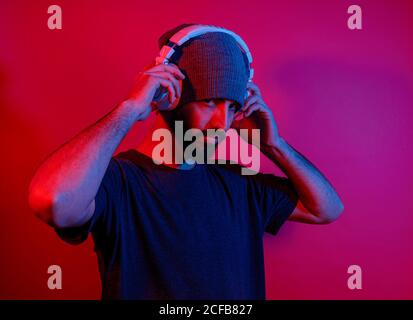 Stylish bearded guy in hat and headphones enjoying good music under red neon light looking at camera Stock Photo