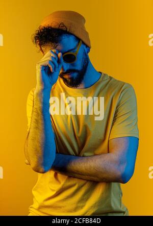 Serious thoughtful bearded male in stylish sunglasses and hat touching face while standing under bright yellow light Stock Photo