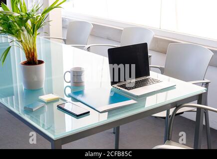 Organized laptop with cellphone and paper folder and mug on glass table with empty chairs around in modern office on daytime Stock Photo