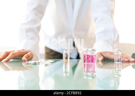 Crop male in lab coat leaning on glass table with arranged vials and medicines for carrying out scientific experiment Stock Photo