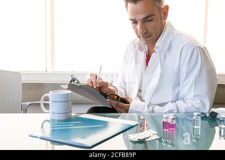 Laboratory assistant sitting at glass table with samples and pills taking notes on clipboard and looking down Stock Photo