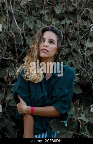 Stylish curly haired lady tourist in dark green shirt and bodysuit looking away while walking on forest with fig tree on background Stock Photo