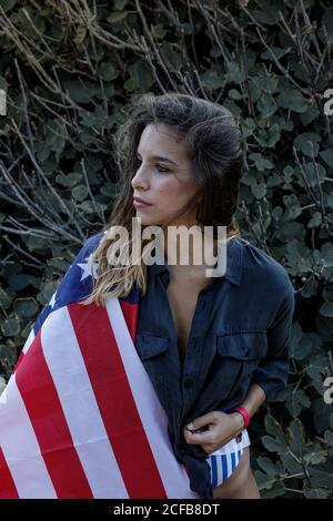 Cheerful curly haired female hipster in casual black shirt and American flag on back looking away with green plants on background Stock Photo