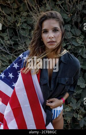 Cheerful curly haired female hipster in casual black shirt and American flag on back looking at camera with green plants on background Stock Photo